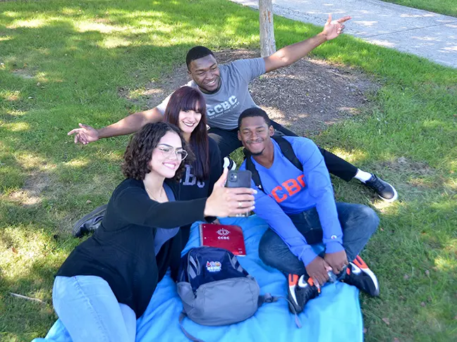 A group of students outside taking a selfie together on a picnic blanket.