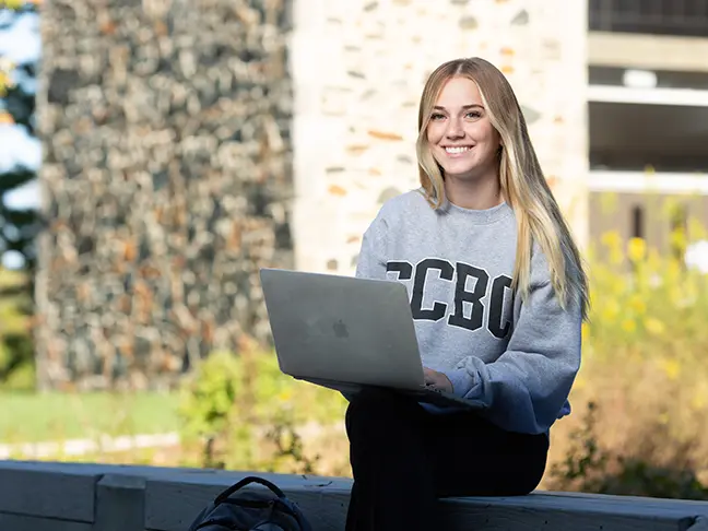 A woman student in a CCBC sweatshirt works on her laptop outside