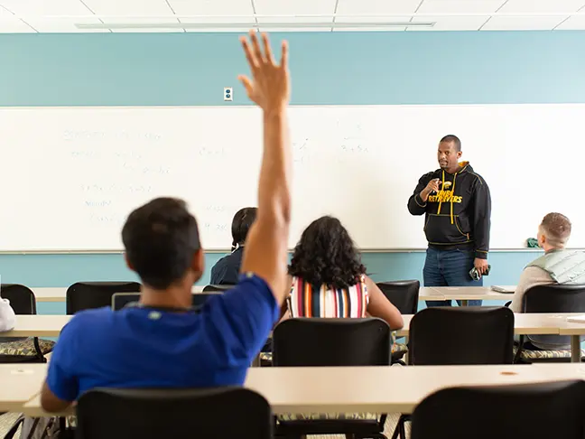 A student raises his hand in a classroom full of students as a teacher calls on him