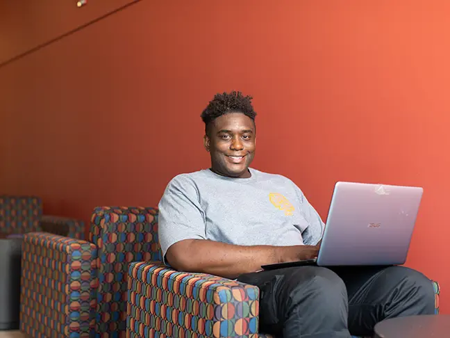 A student sits on an armchair with his laptop, smiling