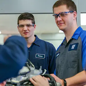 Two mechanic students observe a professional.